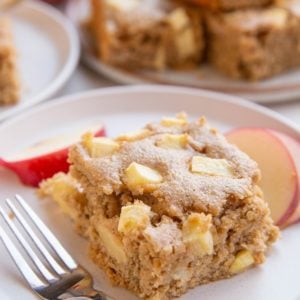 Slice of applesauce cake on a white plate with a wood inlay fork to the side, fresh apple slices all around and a plate with the rest of the cake in the background.