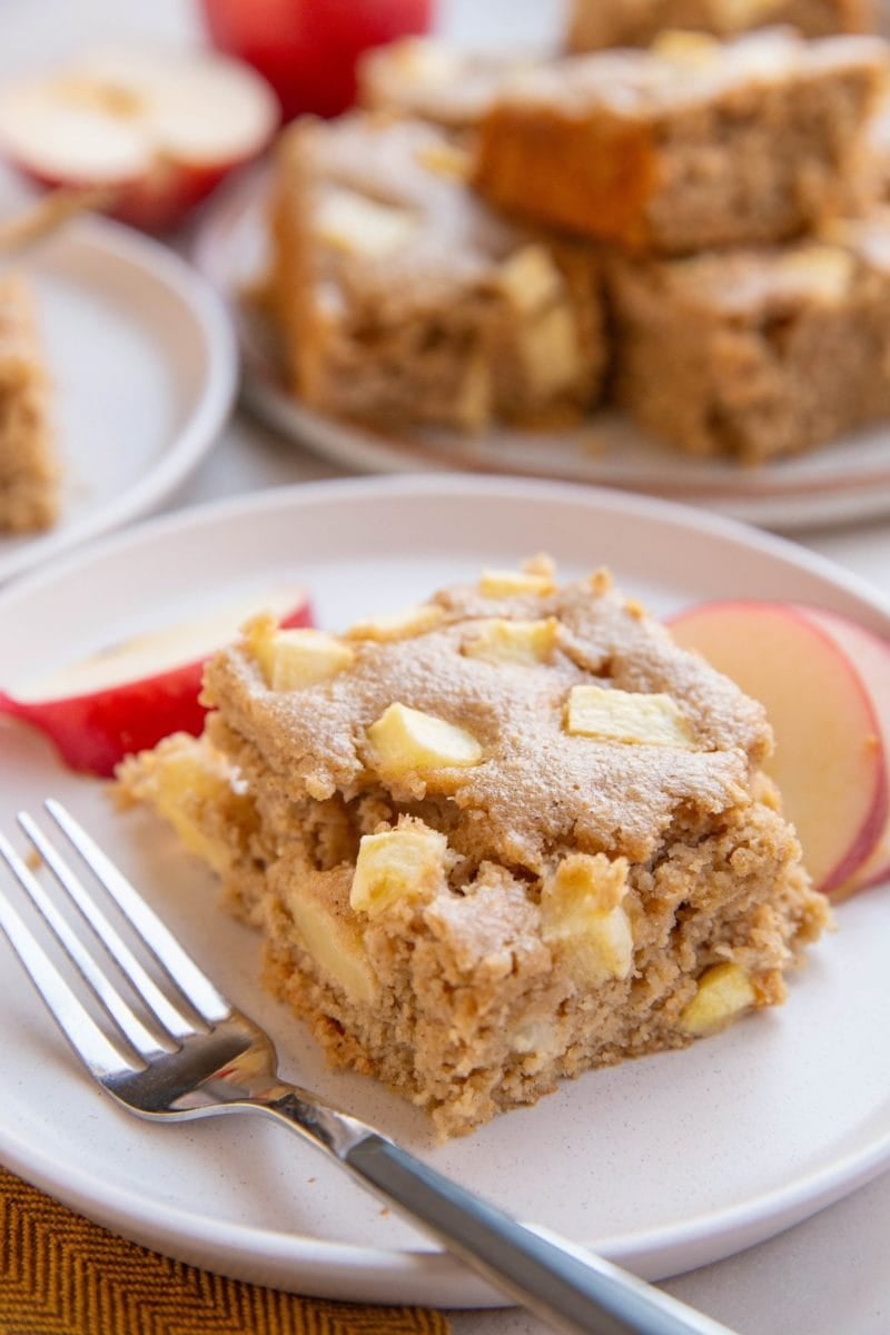 Slice of applesauce cake on a white plate with a wood inlay fork to the side, fresh apple slices all around and a plate with the rest of the cake in the background.