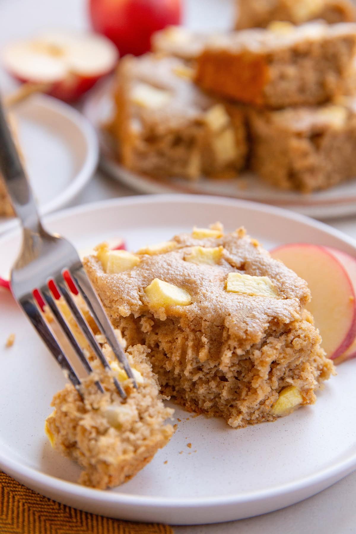 Slice of applesauce cake with a fork taking a bite out and sliced apples to the side.