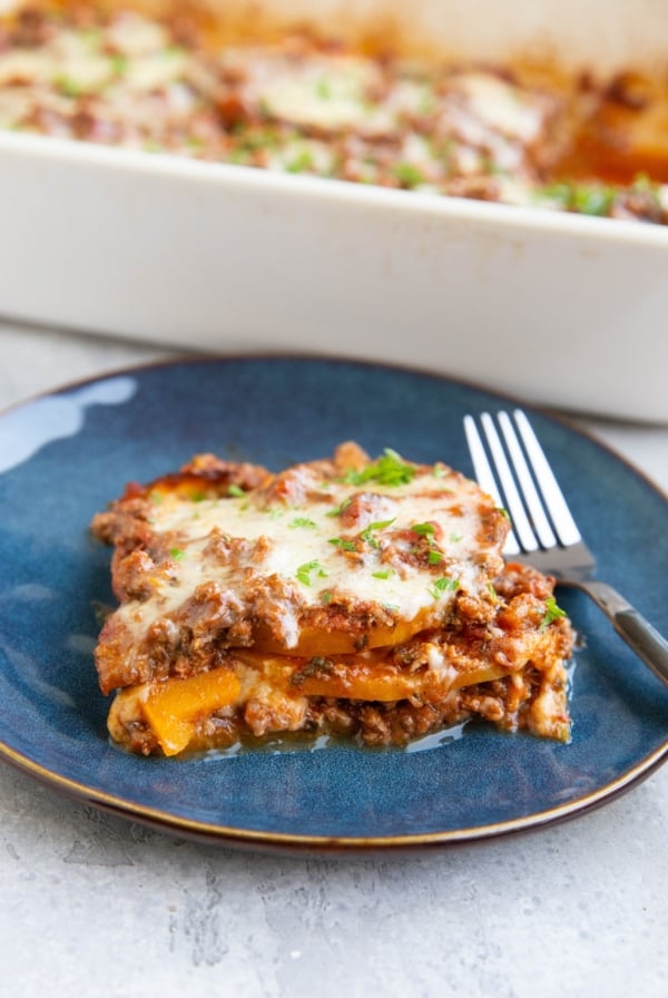 Slice of butternut squash lasagna on a blue plate with the rest of the pan of lasagna in the background.
