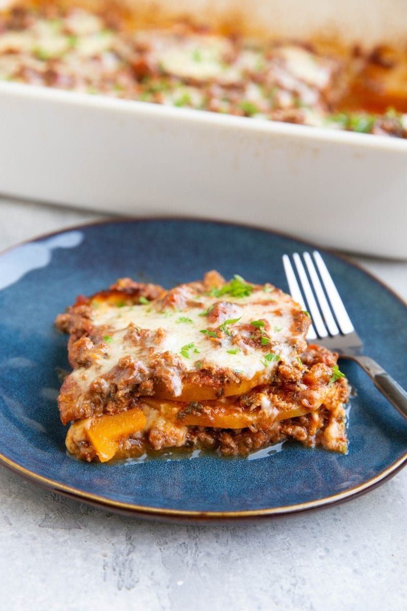 Slice of butternut squash lasagna on a blue plate with the rest of the pan of lasagna in the background.