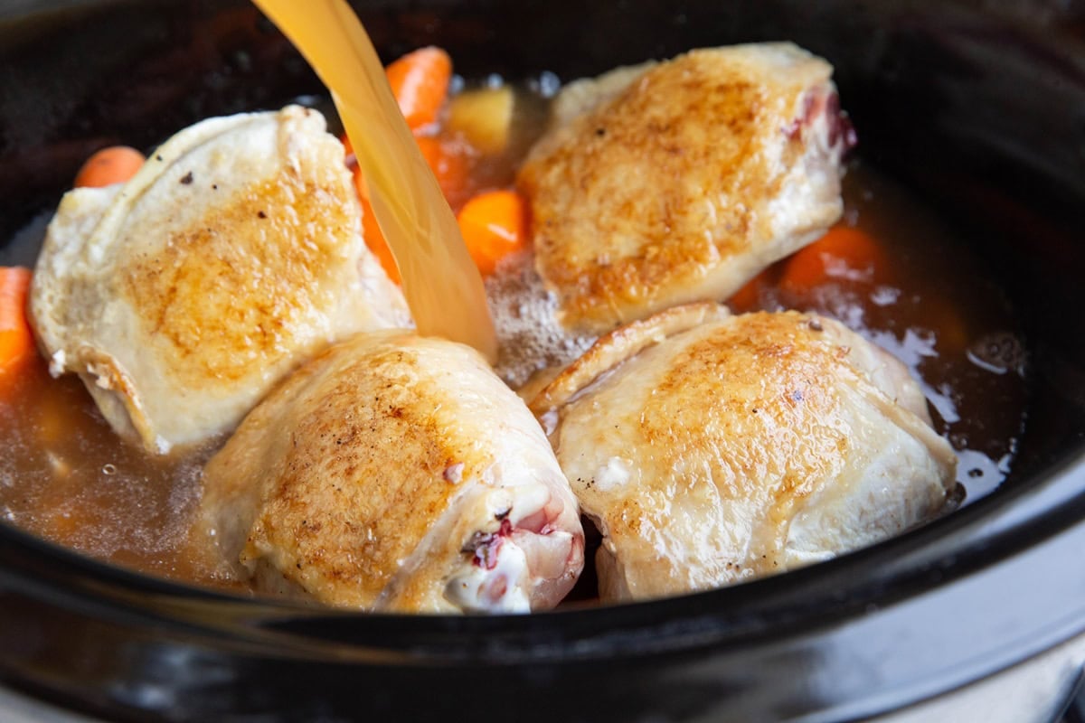 Pouring chicken broth into a slow cooker with chicken and vegetables to make chicken stew.