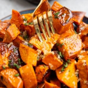 Roasted sweet potatoes on a plate, coated with butter, honey and garlic, with a gold fork picking one up.