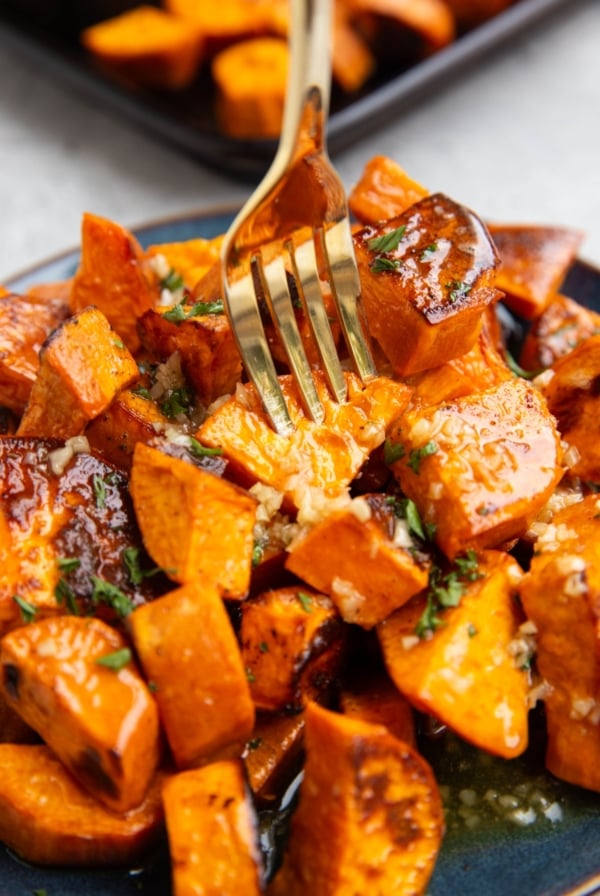 Roasted sweet potatoes on a plate, coated with butter, honey and garlic, with a gold fork picking one up.