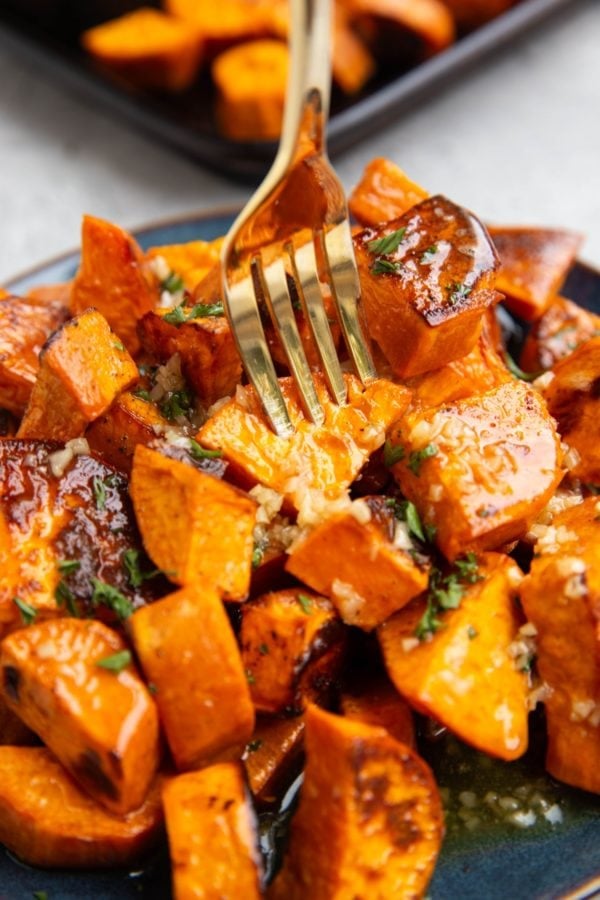 Roasted sweet potatoes on a plate, coated with butter, honey and garlic, with a gold fork picking one up.