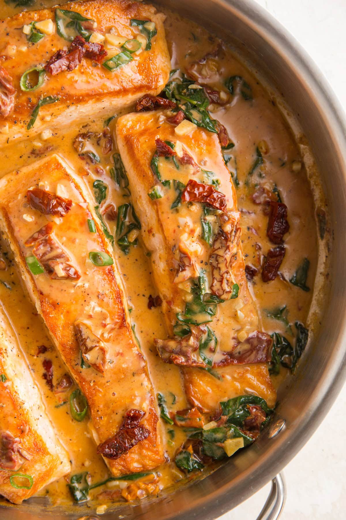 Stainless steel skillet with four salmon fillets in sun-dried tomato sauce.