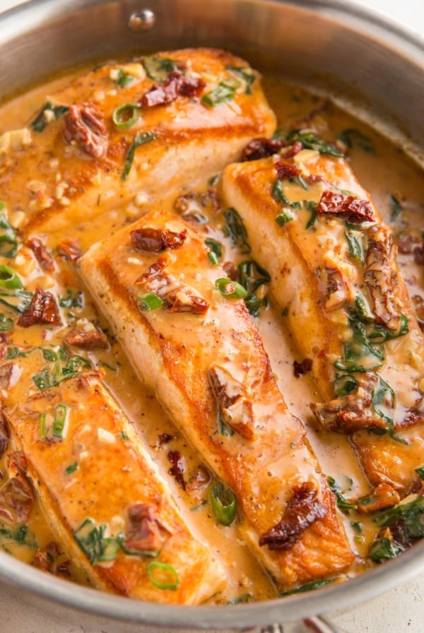 Four cooked salmon filets in a stainless steel skillet with creamy sun-dried tomato sauce. Ready to serve.