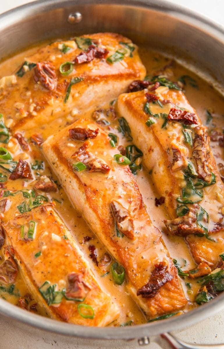 Four cooked salmon filets in a stainless steel skillet with creamy sun-dried tomato sauce. Ready to serve.