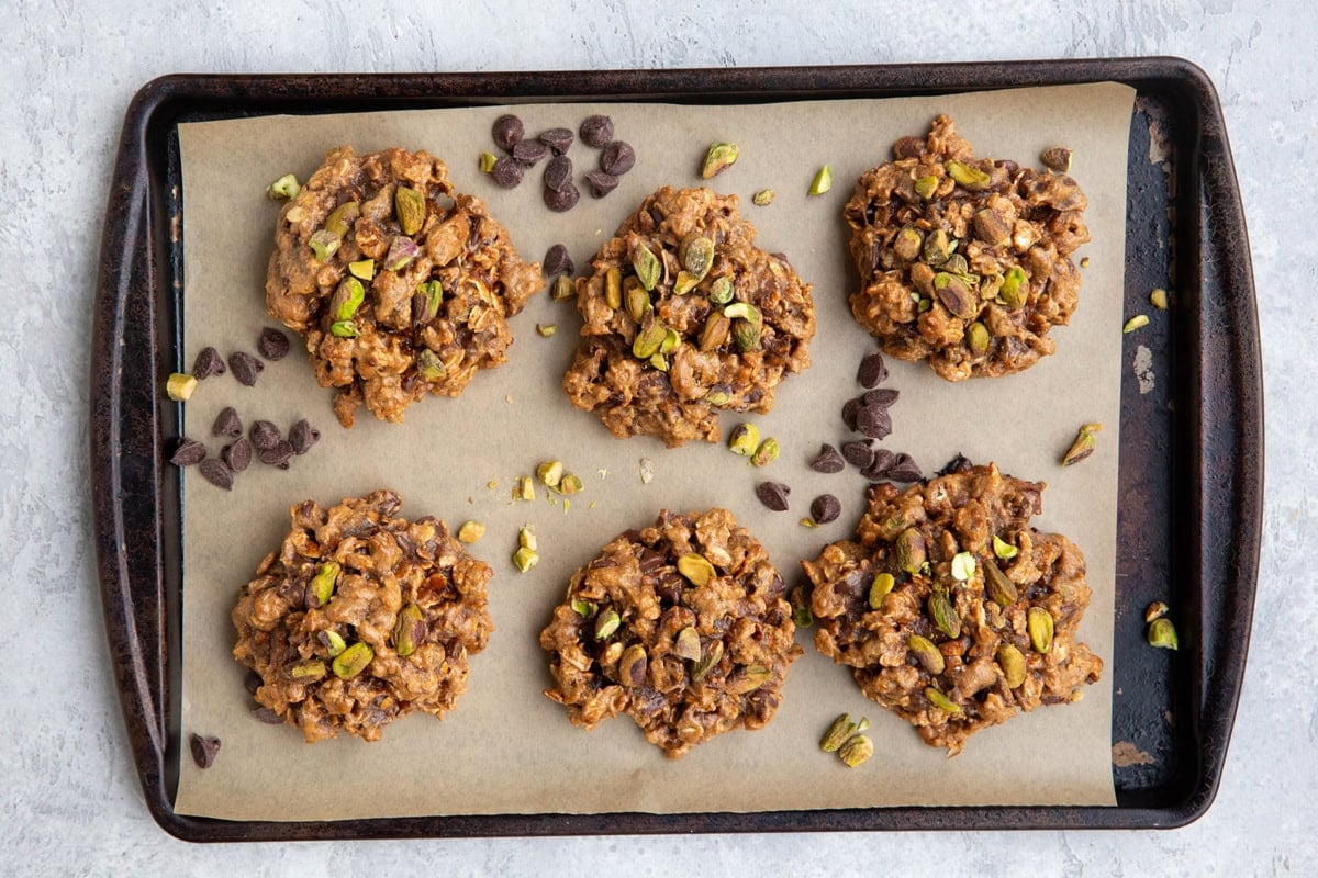 Six large pistachio cookies on a baking sheet with chocolate chips and chopped pistachios sprinkled all around.