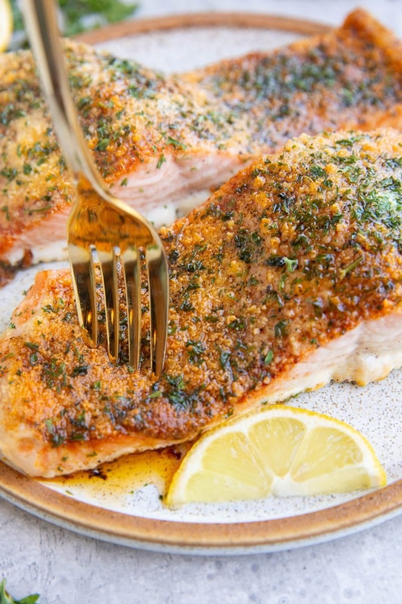 Two parmesan crusted salmon filets on a plate with a fork taking a bite and a lemon wedge to the side.