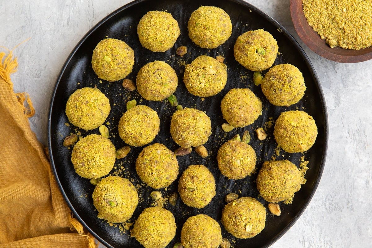 Pistachio energy balls on a black plate with pistachios sprinkled around and a small wooden bowl full of ground up pistachios.