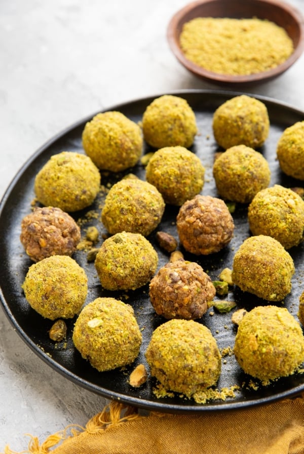 Pistachio energy balls on a black plate with chopped pistachios sprinkled all around. A wooden bowl of ground up pistachios in the background and a golden napkin.