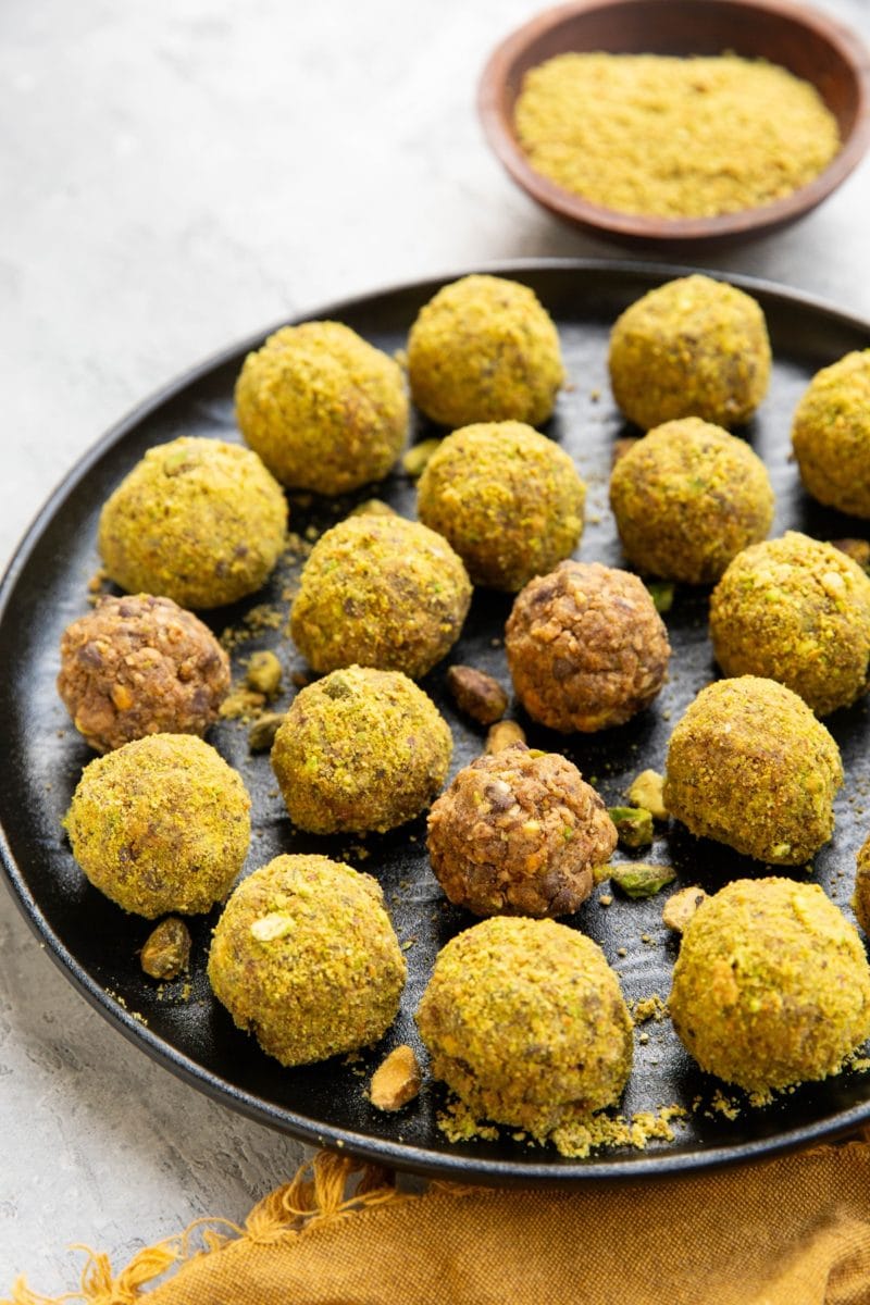 Pistachio energy balls on a black plate with chopped pistachios sprinkled all around. A wooden bowl of ground up pistachios in the background and a golden napkin.