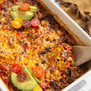 Chicken Taco Casserole with a wooden spoon scooping some of the casserole. Sliced avocado and chopped tomatoes top the casserole and there's a golden napkin to the side.