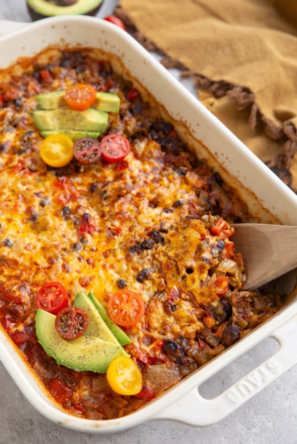 Chicken Taco Casserole with a wooden spoon scooping some of the casserole. Sliced avocado and chopped tomatoes top the casserole and there's a golden napkin to the side.