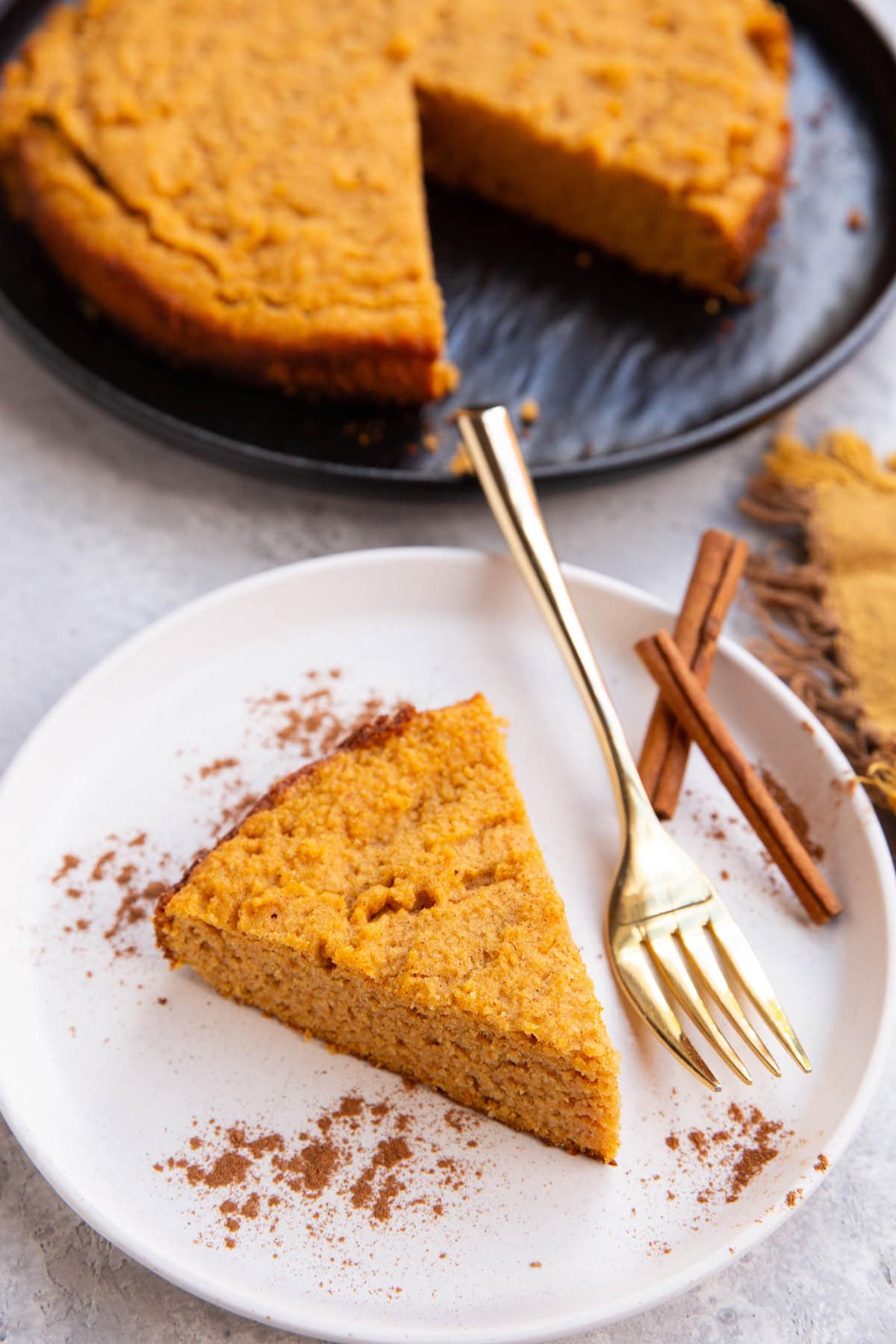 Slice of pumpkin cake on a white plate with the rest of the cake on a black plate in the background.
