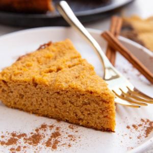 Pumpkin cake on a white plate with pumpkin spice sprinkled all around with a gold fork ready to eat and the rest of the cake in the background.