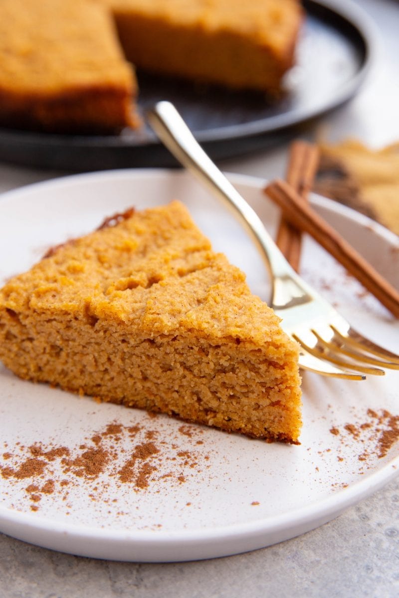 Pumpkin cake on a white plate with pumpkin spice sprinkled all around with a gold fork ready to eat and the rest of the cake in the background.