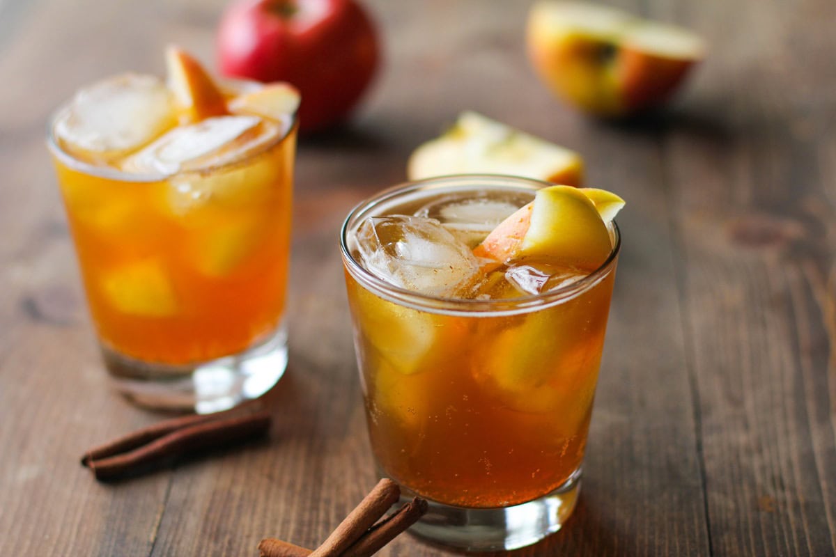 Two apple cider cocktails on a wooden table with fresh apples in the background.