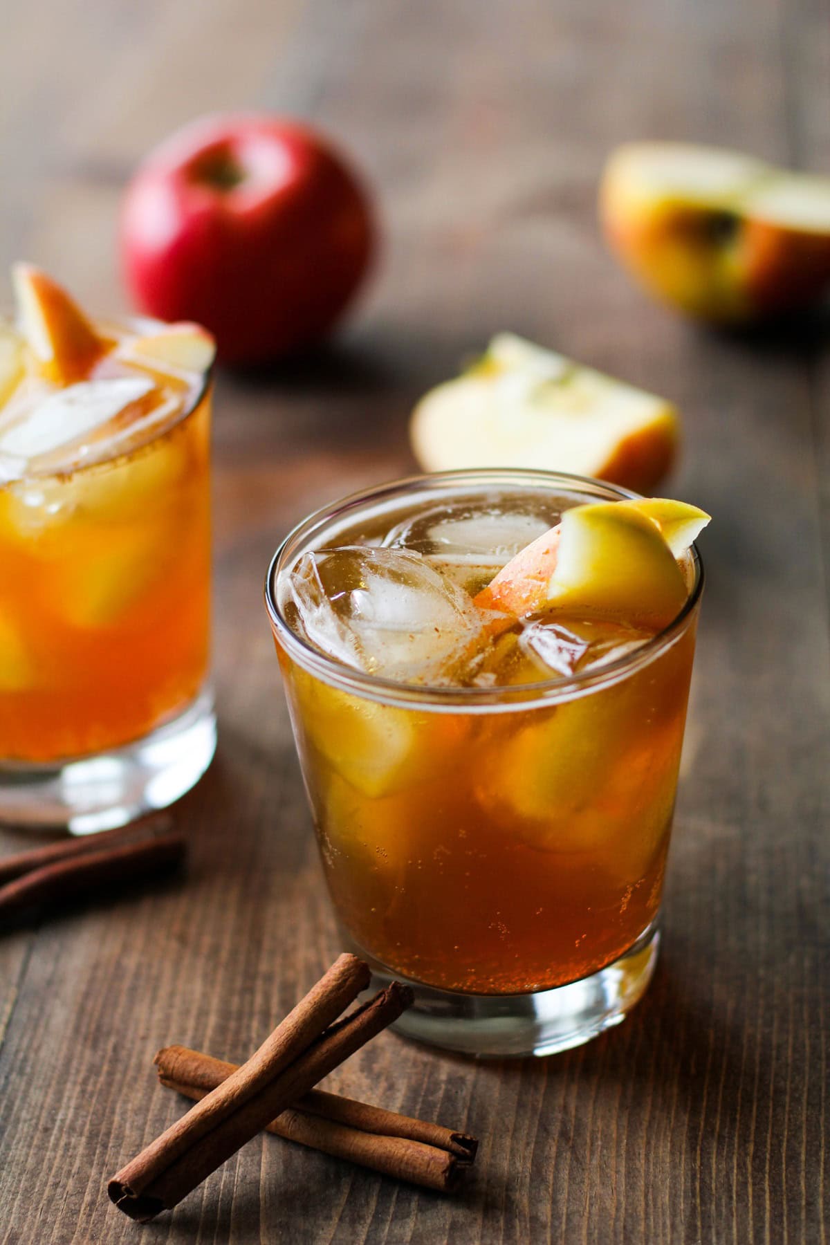 Two apple cider cocktails on a wooden table with cinnamon sticks and apple slices for garnish.