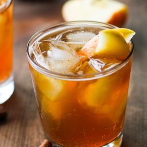 Apple cider cocktail in a cocktail glass with ice and sliced apples with cinnamon sticks to the side and fresh apples in the background.