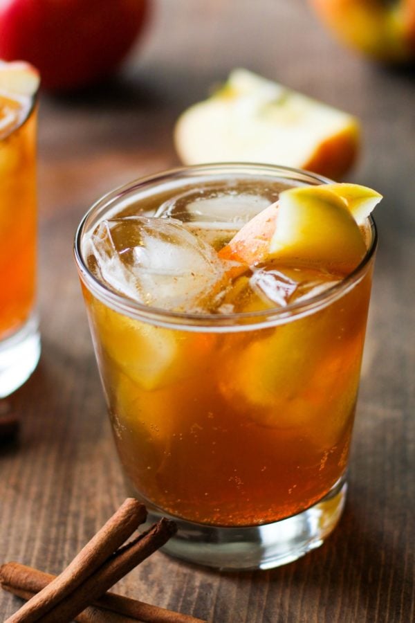 Apple cider cocktail in a cocktail glass with ice and sliced apples with cinnamon sticks to the side and fresh apples in the background.