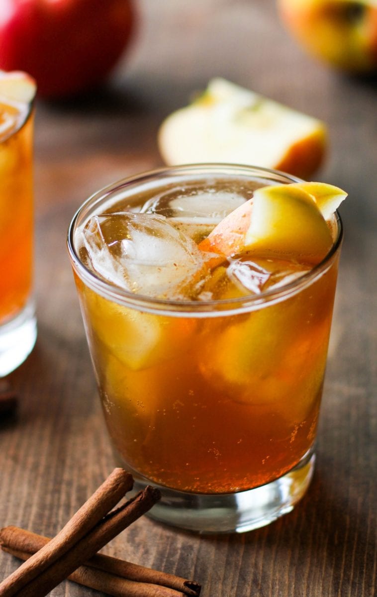 Apple cider cocktail in a cocktail glass with ice and sliced apples with cinnamon sticks to the side and fresh apples in the background.