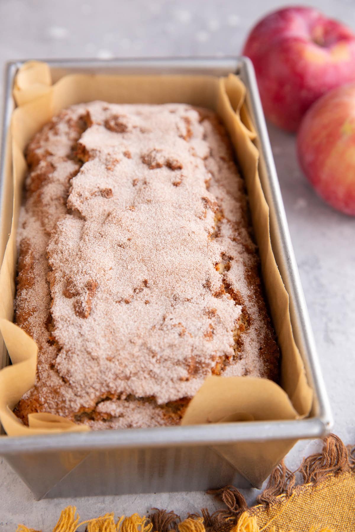 Loaf pan with apple cider donut bread sprinkled with cinnamon and sugar and two fresh apples to the side.