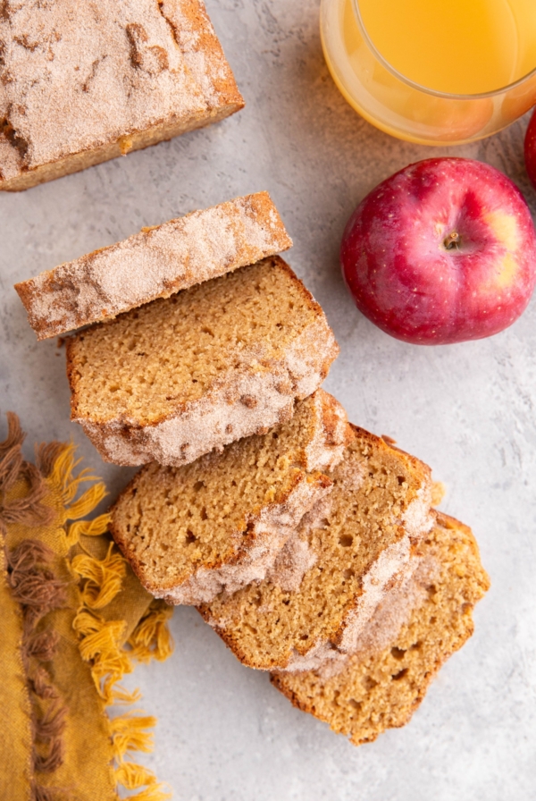 Loaf of apple cider bread cut into slices with a fresh apple and a glass of apple cider to the side.