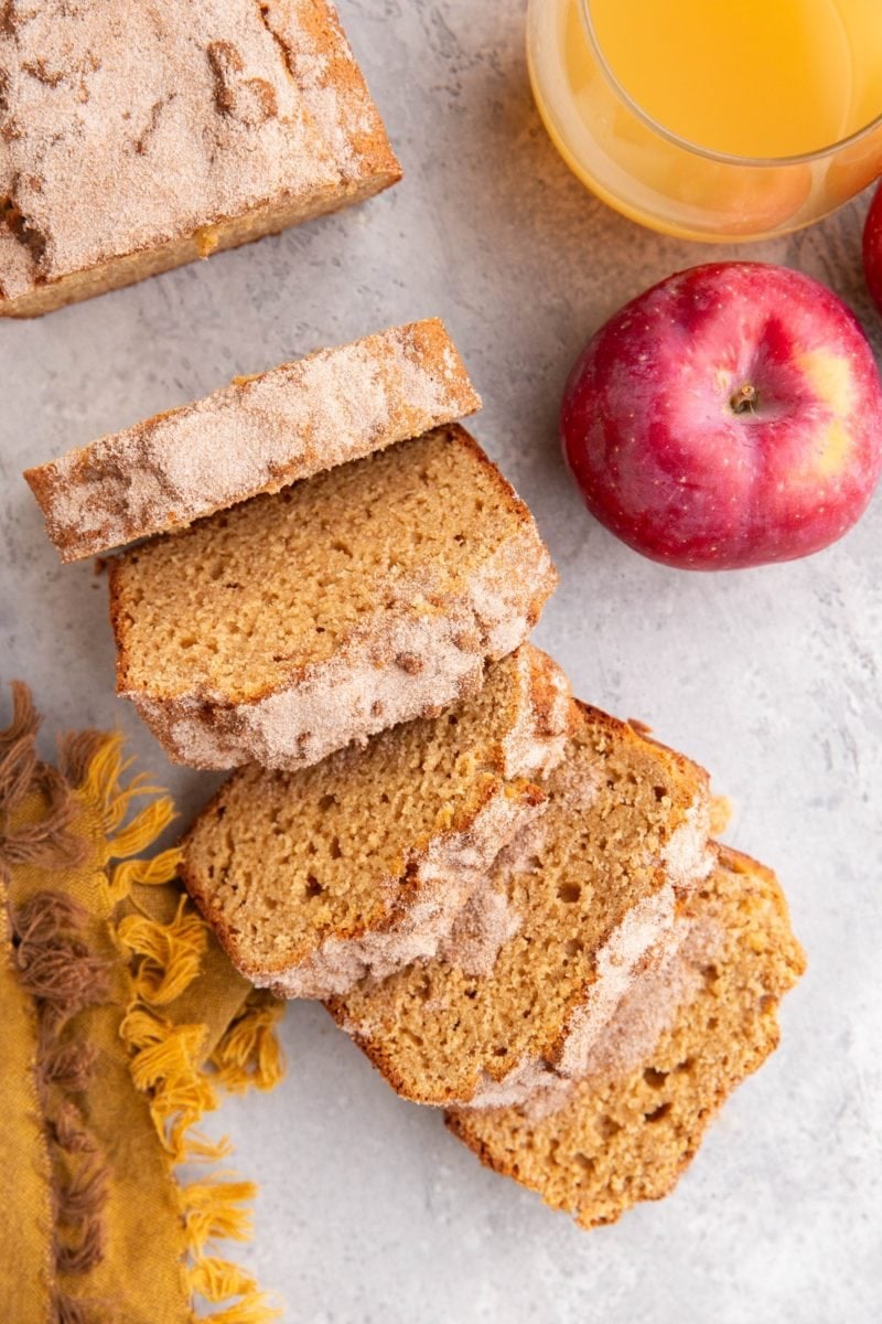 Loaf of apple cider bread cut into slices with a fresh apple and a glass of apple cider to the side.
