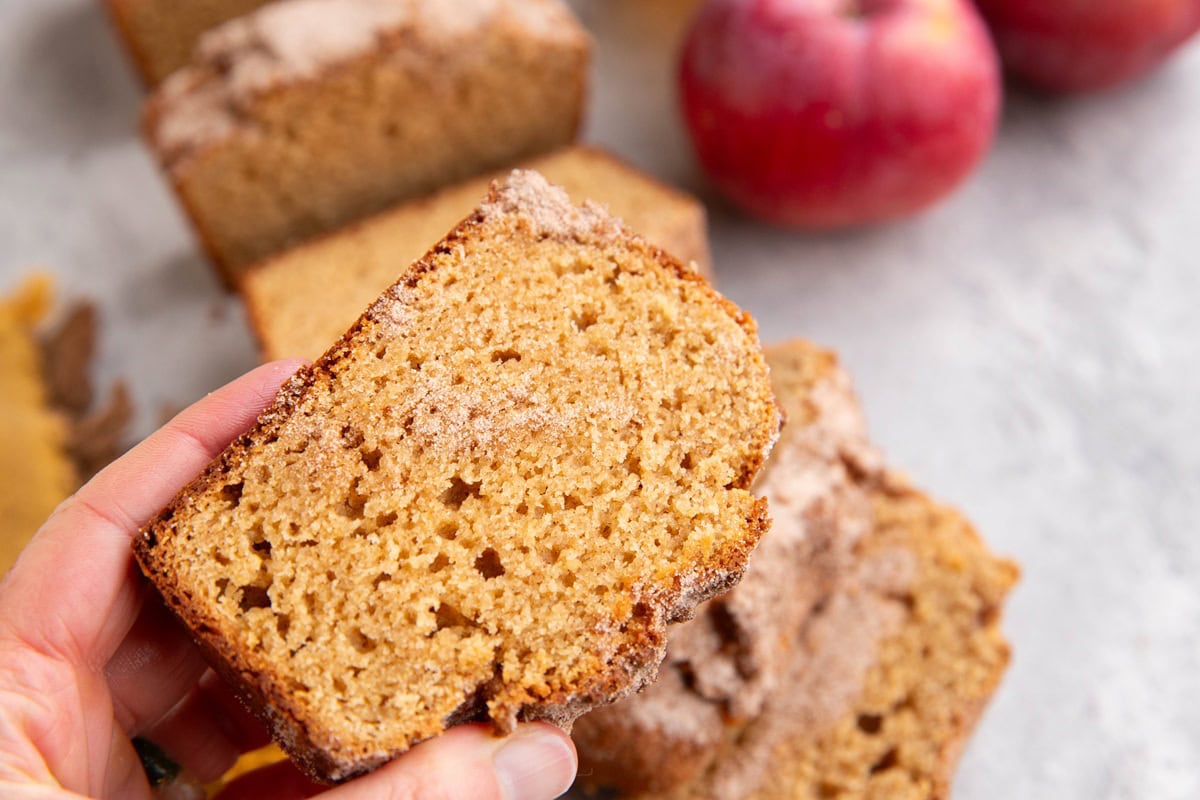 Hand holding a slice of apple cider bread with the rest of the loaf of bread in the background.