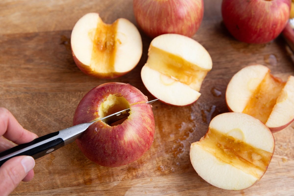 Apples sliced in half on a wooden cutting board with a hand with a paring knife cutting a fresh apple in half.