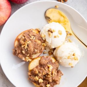 White bowl with two cobbler stuffed apples with two scoops of vanilla ice cream to the side. Fresh apples and walnuts to the side of the bowl and a golden spoon and golden napkin.