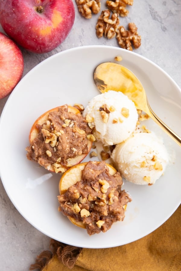 White bowl with two cobbler stuffed apples with two scoops of vanilla ice cream to the side. Fresh apples and walnuts to the side of the bowl and a golden spoon and golden napkin.