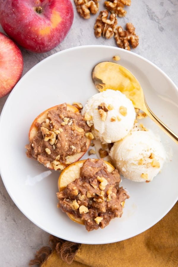 White bowl with two cobbler stuffed apples with two scoops of vanilla ice cream to the side. Fresh apples and walnuts to the side of the bowl and a golden spoon and golden napkin.