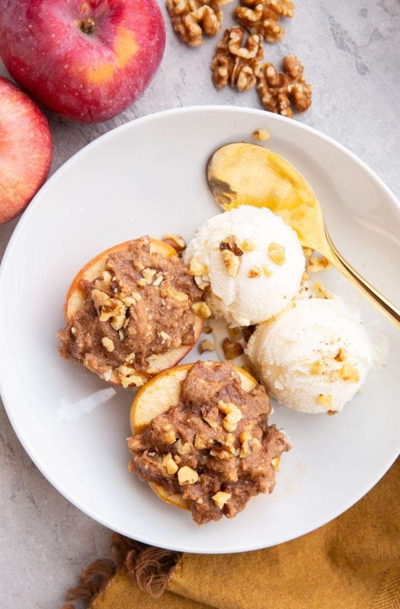 White bowl with two cobbler stuffed apples with two scoops of vanilla ice cream to the side. Fresh apples and walnuts to the side of the bowl and a golden spoon and golden napkin.