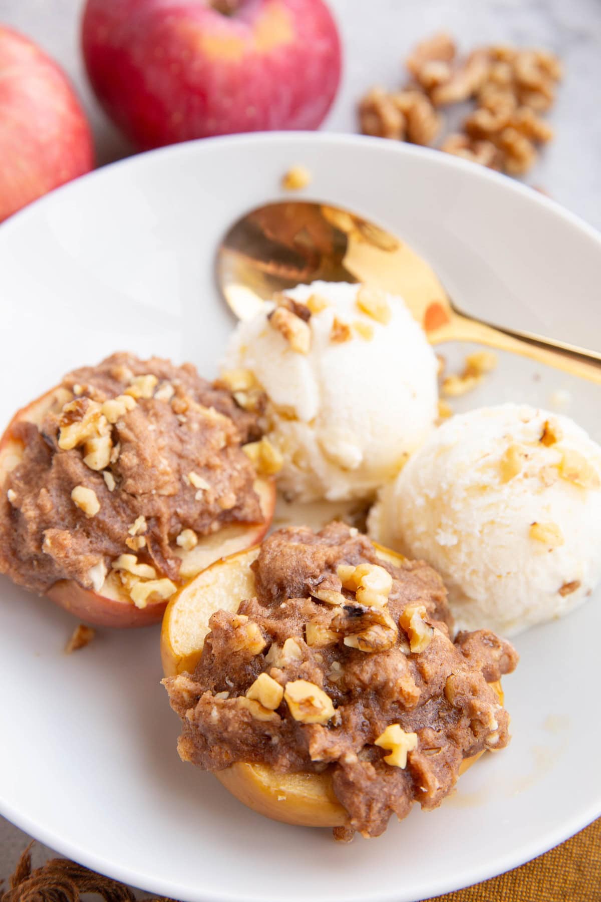 Two stuffed baked apples in a bowl with vanilla ice cream, sprinkled with walnuts and fresh apples in the background.