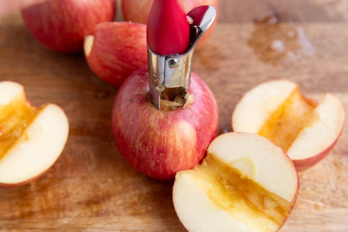 Fresh apples on a wooden cutting board with an apple corer removing the core from one of the apples.