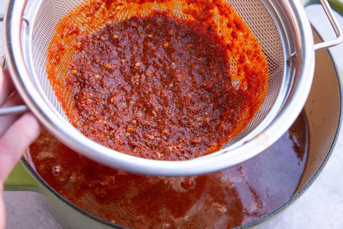 Strainer with chili pulp inside, straining chili sauce into a large pot.
