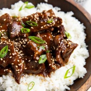 Wooden bowl full of Mongolian beef on top of white rice, sprinkled with sesame seeds and green onions.