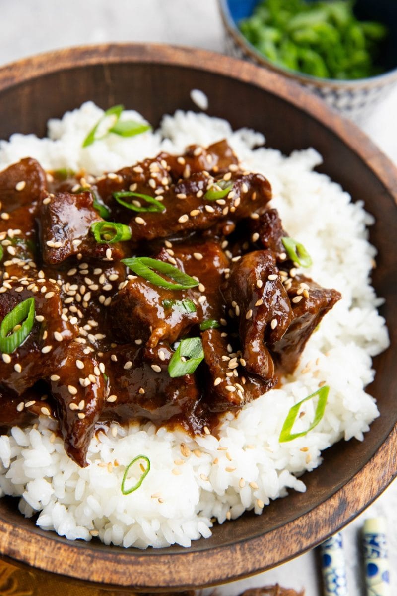 Wooden bowl full of Mongolian beef on top of white rice, sprinkled with sesame seeds and green onions.