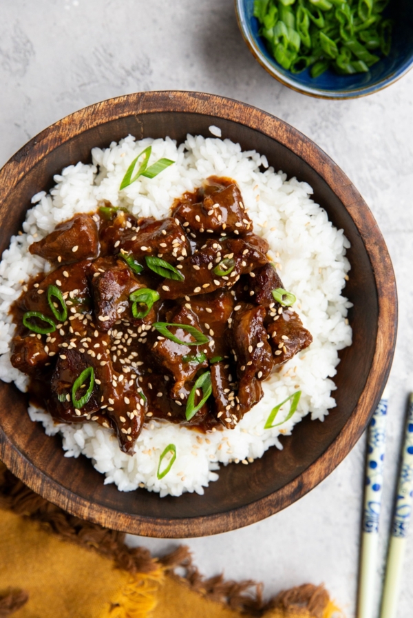 Easy Crock Pot Mongolian Beef in a wooden bowl on top of white rice, sprinkled with sesame seeds and green onions. Chop sticks and a napkin to the side and a bowl of green onions.