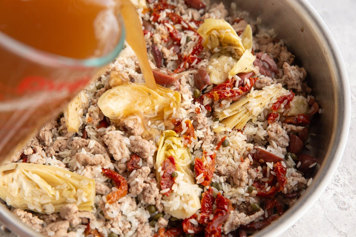 Pouring chicken broth into a skillet with rice, artichoke hearts, sun-dried tomatoes, ground turkey, and olives.