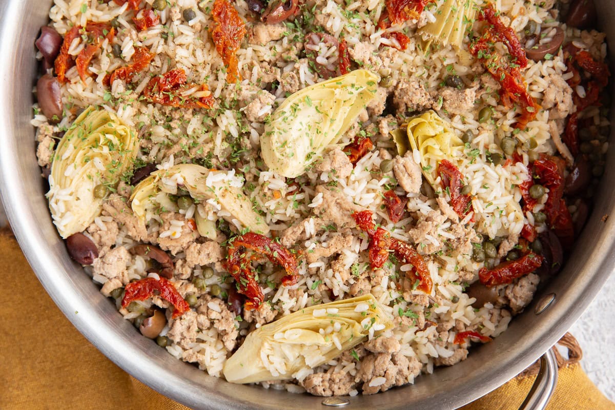 Stainless steel skillet with ground turkey, rice, artichoke hearts, capers, and kalamata olives. Sprinkled with dried parsley.