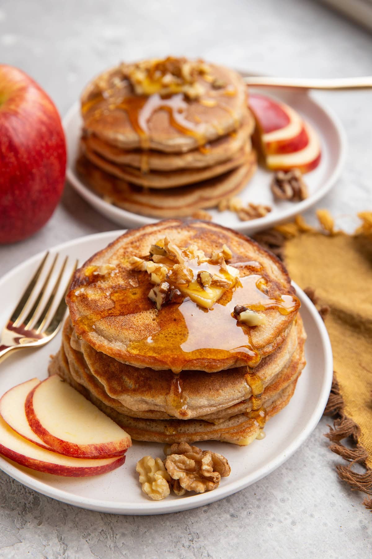 Two plates with stacks of apple pancakes with chopped walnuts on top and sliced apples to the side. A fresh apple and a golden napkin to the side.