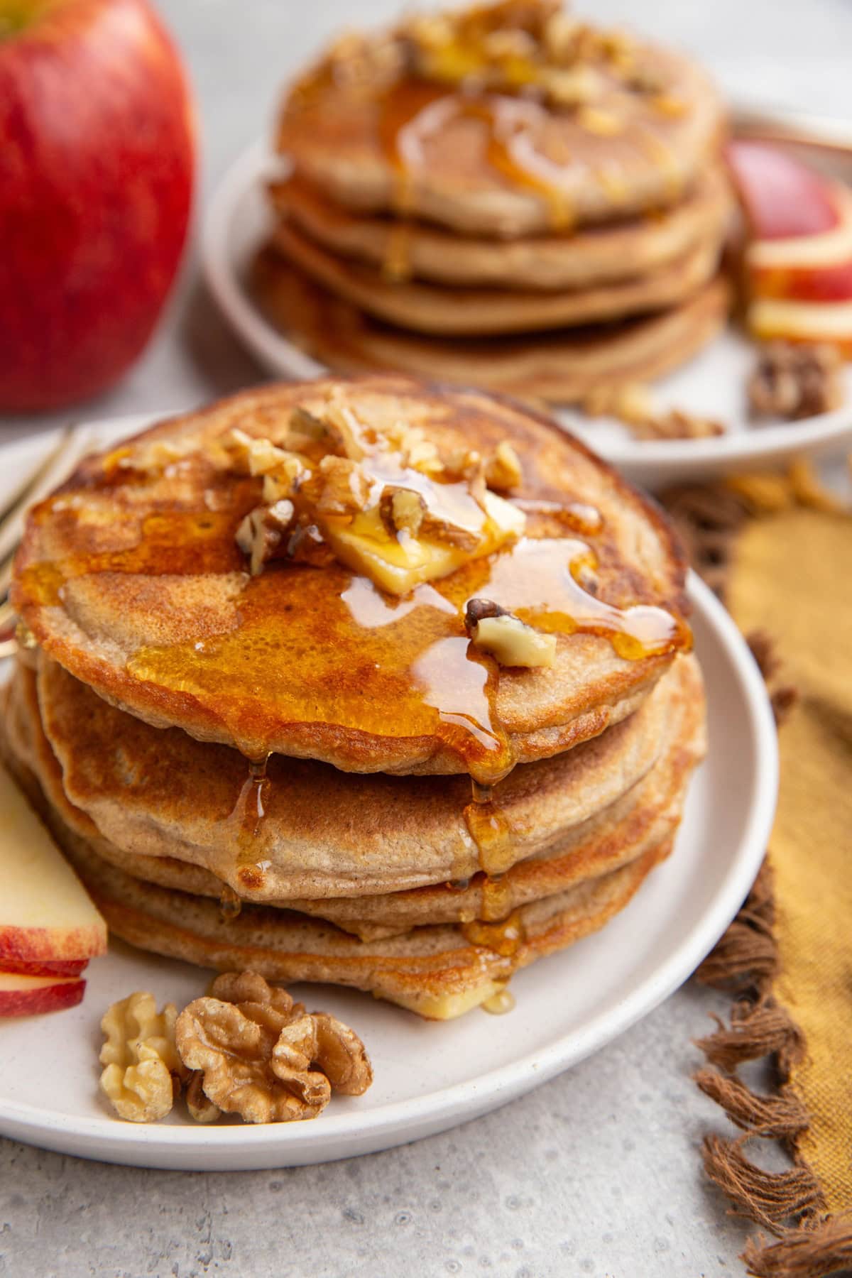Stack of apple pancakes on a white plate with another stack of pancakes in the background. Pancakes are topped with butter, chopped walnuts and honey. Raw walnuts and sliced apple to the side.