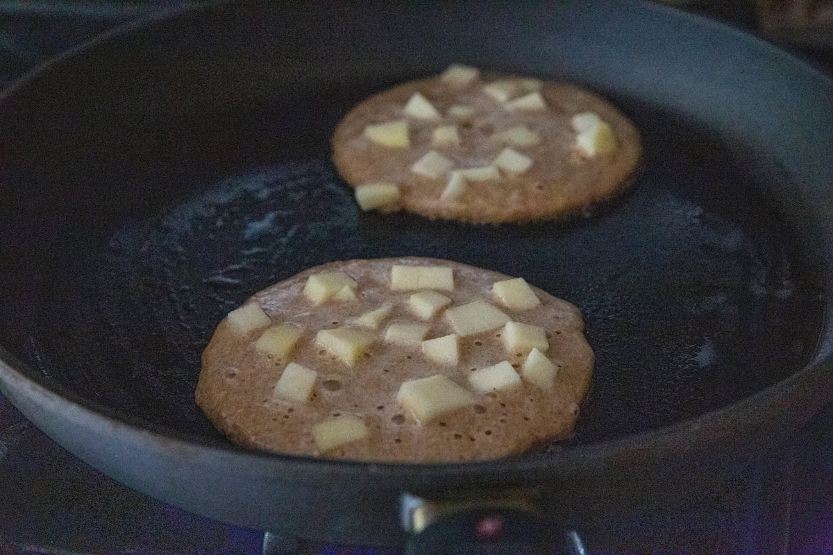 Skillet with two apple pancakes cooking in it with chopped apples on top and little air bubbles rising to the top of the pancake batter.