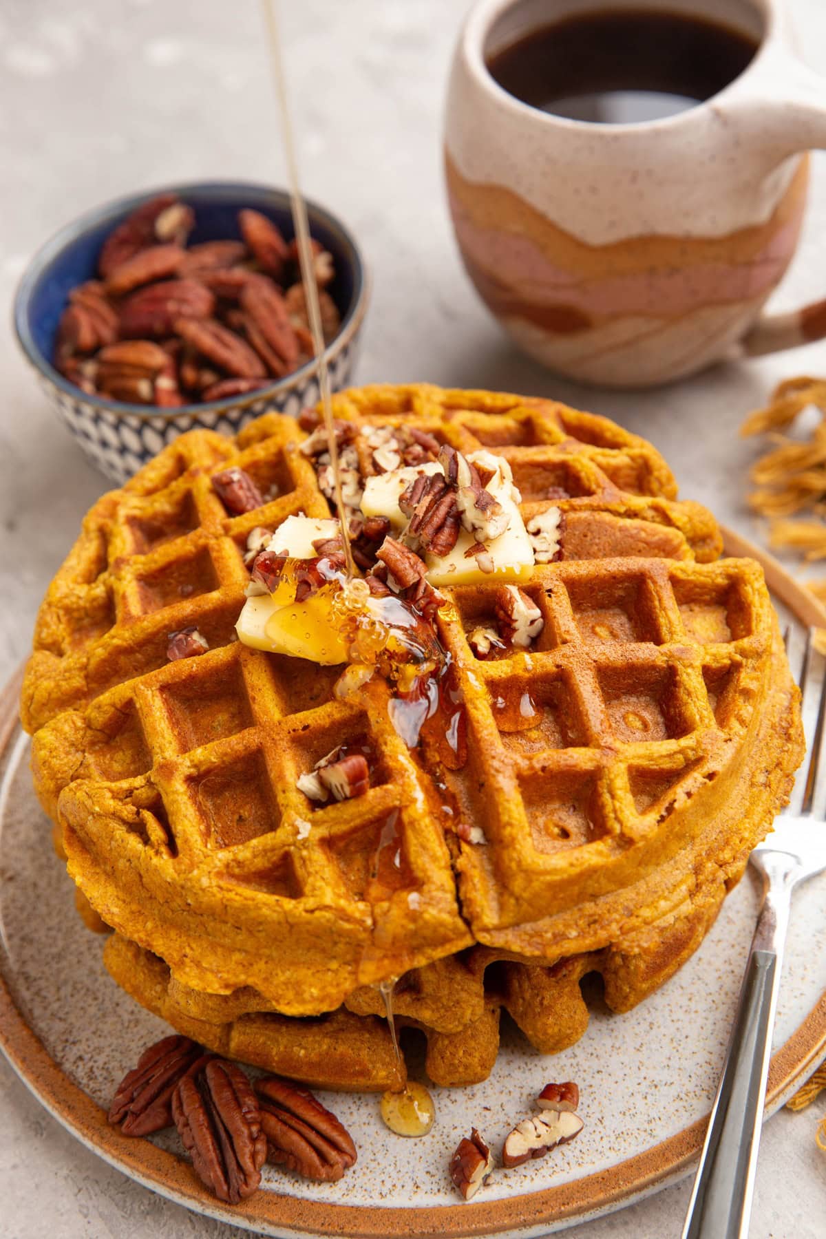 Stack of pumpkin waffles with honey being drizzled on top with butter and chopped pecans. A cup of coffee and a bowl of pecans in the background.