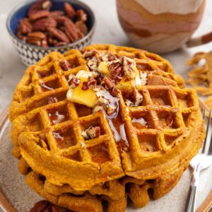Stack of three pumpkin waffles on a plate with butter, honey, and chopped pecans on top. A mug of coffee and pecans in the background.