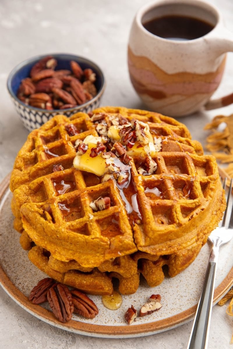 Stack of three pumpkin waffles on a plate with butter, honey, and chopped pecans on top. A mug of coffee and pecans in the background.